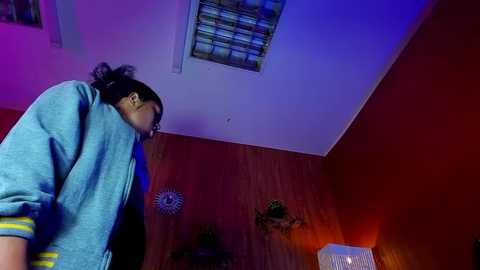 Media: Video of a young person with dark hair and glasses, wearing a blue hoodie, leaning forward in a dimly lit room with red walls and blue lighting, looking down.