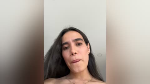 Media: Video of a light-skinned transgender woman with long, dark hair, wearing a necklace, standing against a plain white wall. Her expression is neutral.