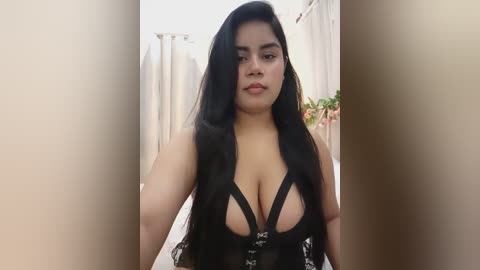 Media: Video of a young woman with long, straight black hair, wearing a black lace lingerie top that reveals ample cleavage, standing indoors near a white curtain.