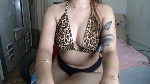 Media: Video of a woman with light skin and long red hair, wearing a leopard-print halter bikini top, black bikini bottoms, and a gold bracelet. She sits on a bed with a teddy bear and pink bedding in the background.