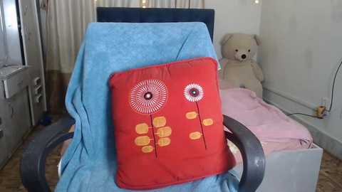 Media: A video of a child's bedroom with a blue chair covered in a red pillow featuring yellow flower designs, next to a teddy bear, a pink blanket, and a white wall.