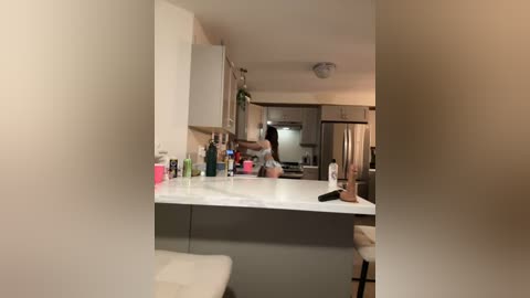 Media: Video of a woman in a kitchen, mid-jump, wearing a black tank top and shorts, with a blurred background, showcasing modern appliances and white cabinets.