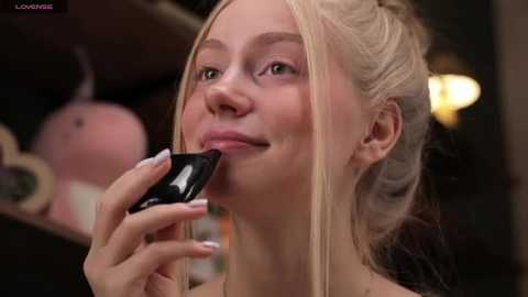 Media: Video of a fair-skinned young woman with blonde hair, styled in a messy bun, applying lipstick with a black lipstick brush. Background is blurred, with indistinct shapes and soft lighting.