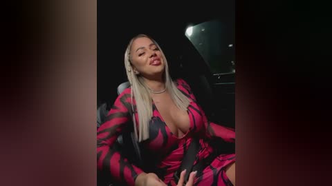 Media: Video of a curvy, light-skinned, blonde woman with long hair, wearing a low-cut, red and black striped dress, sitting in a dimly-lit car, with a shadowy background.