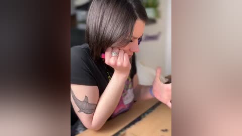 Media: Video of a woman with shoulder-length dark hair, wearing a black shirt and pink apron, leaning on a wooden table, with a tattoo on her left arm.