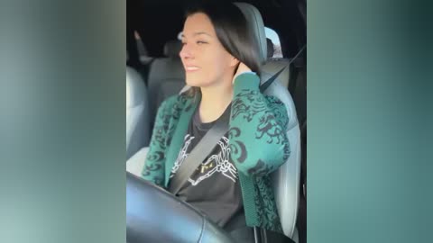 Media: Video of a woman with medium skin tone, dark hair, and a light blue jacket with intricate black designs, seated in a car. She has a relaxed expression, wearing a black graphic t-shirt. The car interior is modern with gray seats.