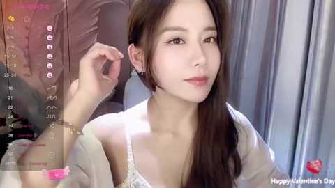 Media: Video of a young Asian woman with fair skin, long dark hair, and brown eyes, wearing a white lace bra, holding a phone, in a dimly lit room.