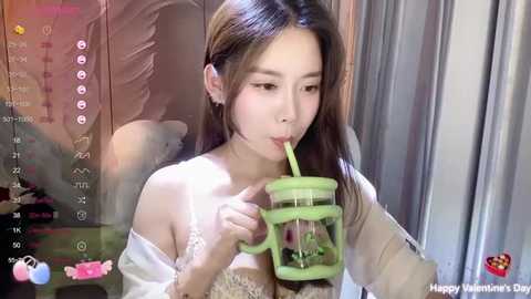 Media: Video of an East Asian woman with long, straight brown hair, drinking from a green, heart-shaped straw, wearing a white off-shoulder top. Background features a pink panda plush toy and gray curtains.