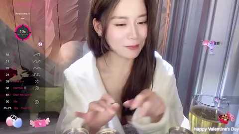 Media: A video of a young Asian woman with long brown hair, wearing a white robe, smiling while holding a pink candy heart in a dimly lit room with a glass of wine and a glass of water.