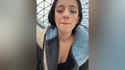 Media: Video of a young woman with medium skin tone, straight black hair, wearing a black jacket with a light blue lining, and a black top, standing in front of a modern glass and metal structure.