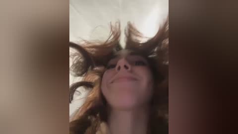 Media: A video captures a young woman lying on her back, with her long, wavy brown hair splayed out, smiling up at the camera. The background is blurred, featuring a beige or light brown wall.