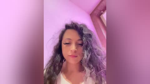 Media: A video of a young woman with long, wavy, gray-streaked hair, wearing a white top, standing against a pink wall. Her expression is calm, eyes closed, and she has a subtle smile.