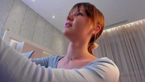 Media: Video of a fair-skinned woman with short, light brown hair, wearing a light blue cardigan, standing in a modern, light-filled room with white walls and a leaf-patterned wallpaper.