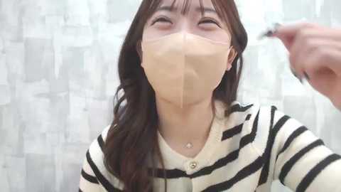 Media: Video of a woman with long brown hair, wearing a beige surgical mask, black-and-white striped sweater, and earrings, in a bathroom with white and gray patterned shower curtain.