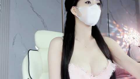 Media: A video of an East Asian woman with long black hair, wearing a white face mask and a lacy, pink bra. She sits in a modern, minimalist room with a white chair and gray marble wall.