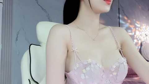 Media: A video of an Asian woman with fair skin, wearing a delicate, light pink lace bra with floral embroidery, sitting on a white chair against a marble wall with a crystal chandelier.