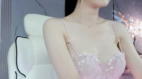 Media: Video of a fair-skinned woman in a pale pink, lace-trimmed bra, with a delicate gold necklace, seated on a white leather chair in a modern, softly lit room with marble walls.