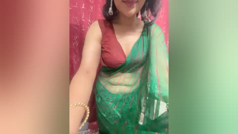 Media: A video of a South Asian woman in a red and green sari with a plunging neckline, wearing gold jewelry and sitting against a floral-patterned pink backdrop.