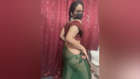 Media: Video of a woman in a red sari with a green drape, wearing a black face mask, standing against a red floral-patterned curtain.