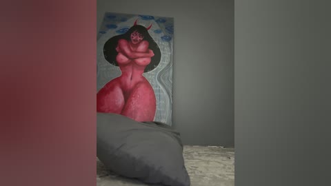 Media: Video of a surreal, red-skinned woman with large breasts and wide hips, wearing a black hat, standing in a gray room with a blue sky mural on the wall and a gray cushion on the floor.