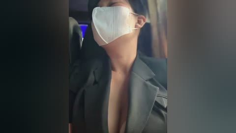 Media: Video of a woman wearing a black suit, white surgical mask, and dark hair, with a blurred background suggesting a dimly lit room.