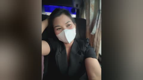 Media: A video of a woman with long black hair, wearing a white face mask, black blazer, and taking a selfie in a dimly lit room.