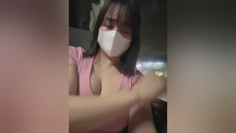 Media: Video of a young Asian woman with light skin, wearing a pink top, white mask, and black hair, sitting in a dimly lit room, her arms crossed, and blurred background.