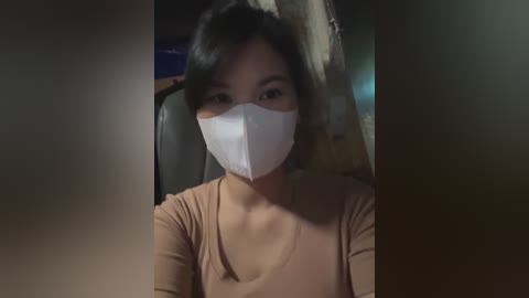 Media: A video of an Asian woman with straight black hair, wearing a white face mask and a beige V-neck shirt, standing indoors with dim lighting, blurred background.