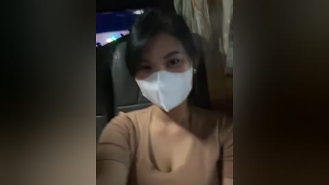 Media: Video of a young East Asian woman with long black hair and medium skin tone, wearing a white face mask and a beige top, indoors with dim lighting.