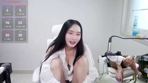 Media: Video of an Asian woman with long black hair, wearing a white blouse, sitting on a chair, smiling. Background features a calendar, desk, and a window with sunlight.