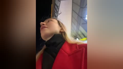 Media: A video shows a young woman with fair skin, long blonde hair, and closed eyes, leaning back against a red background. She wears a black coat, and the setting appears to be a dimly lit, possibly indoor space.