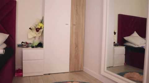 Media: Video of a minimalist bedroom with a white wardrobe, wooden door, white dresser, maroon bed, white pillow, floral arrangement, and a round rug.