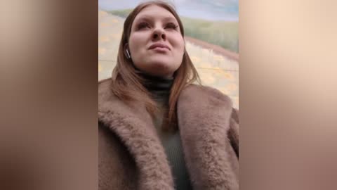 Media: Video of a fair-skinned woman with straight, light brown hair, wearing a brown faux fur coat over a green sweater, standing in front of a painted landscape with green hills and yellow fields.