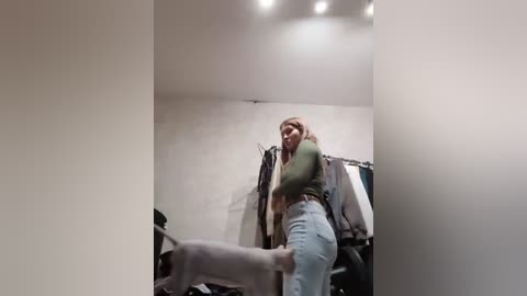 Media: Video of a woman with long blonde hair in a green top and light blue jeans, bending over to pet a dog in a cluttered, dimly lit room with clothes hanging on the wall.