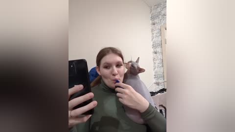 Media: A video of a young woman with light skin and straight brown hair, wearing a green top, taking a selfie with a black smartphone, while a Siamese cat with a blue collar sits behind her.