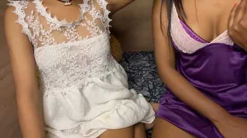 Media: Video of two women in lingerie: one in white lace, the other in purple satin. The background shows a patterned bedspread.