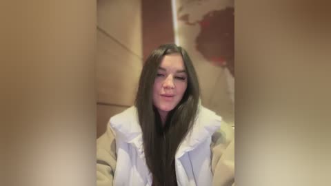 Media: A video of a young woman with long black hair and light skin, wearing a white puffy jacket, standing in a dimly lit, beige-walled bathroom, with blurred, red-painted walls in the background.