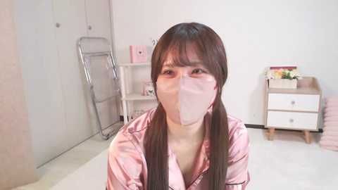 Media: Video of an East Asian woman with long black hair in pigtails, wearing a pink satin robe and a pink surgical mask, sitting on a white floor in a minimalist, white-walled room with a metal step ladder, wooden drawers, and a small table with a flower arrangement.