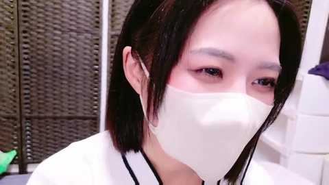 Media: Video of an Asian woman with straight black hair, wearing a white surgical mask, looking to the right. Background features a wicker screen and white tiles.