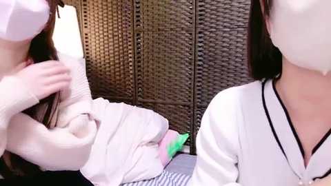 Media: Video of two Asian women in white face masks, one in a white sweater, the other in a white shirt, sitting on a striped bed, against a wicker screen background.