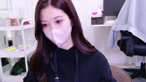 Media: Video of an Asian woman with long brown hair, wearing a black dress and a white surgical mask, standing in a modern office with white walls, a white desk, and a black chair.