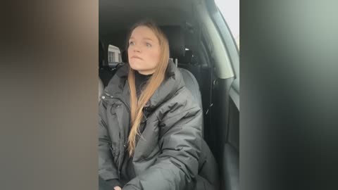 Media: Video of a young Caucasian woman with long, straight blonde hair, wearing a dark, puffy winter jacket, sitting in the back seat of a car, looking out the window with a contemplative expression.