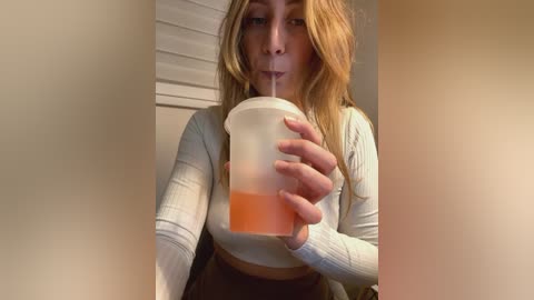 Media: Video of a young woman with shoulder-length blonde hair, wearing a white long-sleeve crop top and black pants, sipping from a white straw in a clear plastic cup. Background includes a white window blind.