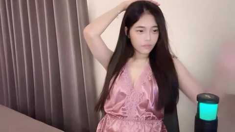 Media: A video of a young East Asian woman with long black hair, wearing a pink satin robe, standing in a dimly lit room with beige walls and a gray curtain.