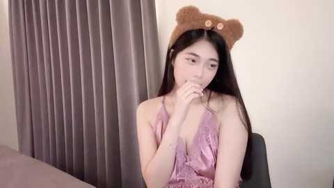 Media: A video of an Asian woman with long black hair, wearing a pink lace camisole and a bear headband, sitting with her fingers to her lips, against gray curtains.