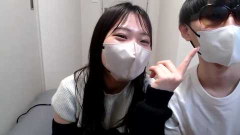 Media: Video of two East Asian women in a white room, wearing white masks and black clothing, with one woman holding a phone to the other's ear.