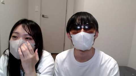 Media: Video of two Asian women wearing white surgical masks, one with long black hair, the other with short black hair, in a sterile, white-walled room with a door in the background.