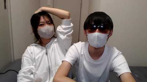 Media: Video of two Asian men in white medical masks, white jackets, and glasses, seated on a grey couch in a sterile, beige-walled room.