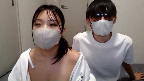 Media: Video of two Asian women in surgical masks, one with a low ponytail, wearing a white jacket, the other with short hair, in a white t-shirt, both seated indoors with plain white walls.