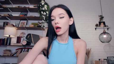 Media: Video of a slim, pale-skinned, long-haired young woman with a nose ring, wearing a light blue halter top, seated in a bookshelf-filled room with white brick walls.